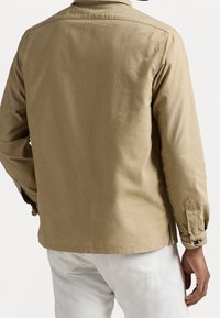 Hnědá košile s dlouhým rukávem a knoflíkovým límcem, má hladký povrch, boční rozparky a knoflíkové manžety, nošena přes světlé kalhoty.