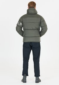 Puffjacka i olivgrönt, med horisontella sömmar, hög krage och en logotyp på vänster arm. Matchas med mörka byxor och stövlar.