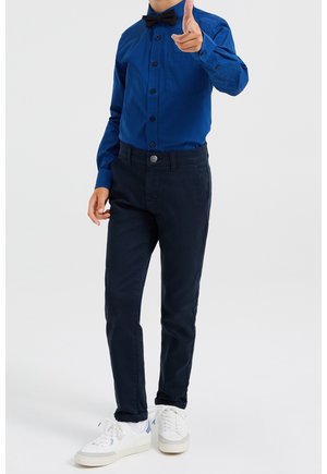 Chłopiec w niebieskiej koszuli w wzory, z czarną muszką i granatowymi spodniami, wskazujący przed siebie prawą ręką, stojący na białym tle.