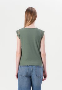 Olivengrønn ermeløs topp med teksturerte blonde detaljer på skuldrene, med rund hals og tettsittende fasong, kombinert med lyseblå jeans.