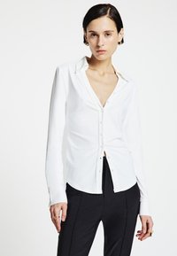 Weißes Langarmhemd mit Kragen und Knopfleiste, aus glattem Stoff. Getragen mit schwarzen, eng anliegenden Hosen, minimalistisch gestaltet.