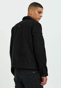 Veste en daim noir avec col, manches longues et poignets à bouton. Présente une étiquette de logo au dos et un ourlet droit. Texture lisse sans motifs.