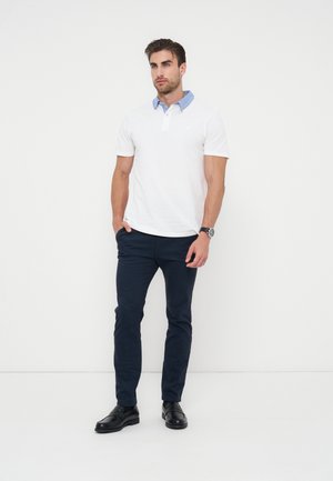 Homme portant un polo blanc avec un col bleu, un pantalon bleu foncé, des chaussures noires et une montre-bracelet, debout devant un fond blanc.