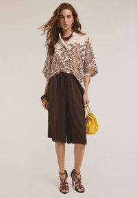 Blusa bianca con motivo floreale marrone, pantaloni corti larghi marroni, tacchi neri con cinturini e una borsa gialla con chiusura a strappo.