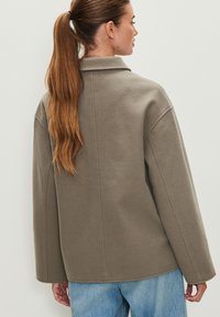 Brūna pārmērīgi liela jaka ar apkakli, izgatavota no mīksta auduma. Piedurknes nolaižas un aizmugurē redzamas redzamas šuves.