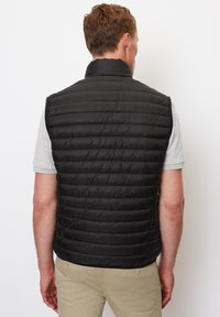 Schwarze Steppweste mit hohem Kragen, gestepptem Muster und ärmellosem Design, getragen über einem hellgrauen T-Shirt mit kurzen Ärmeln.