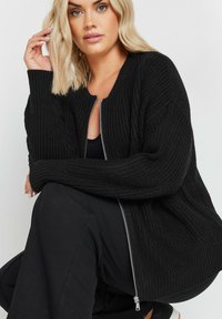 Svart stickad cardigan med ribbad struktur, dragkedja fram och sänkta axlar, bärs över en svart topp och byxor.