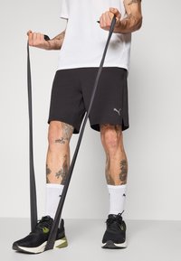 Zwarte sportshorts met zijsplitten, een Puma-logo, gecombineerd met een wit T-shirt, zwarte sneakers en witte sokken. Houdt een weerstandsband vast.