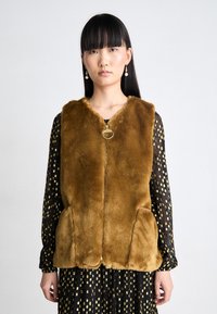 Brauner Kunstpelz-Westen mit V-Ausschnitt, Reißverschluss und zwei vorderen Taschen. Getragen über einem schwarzen Kleid mit goldenen Mustern.