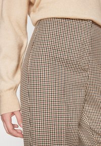 Pantalon à motif pied-de-poule marron et beige avec une coupe ajustée. Tissu texturé présenté à côté d'une manche de pull beige clair.