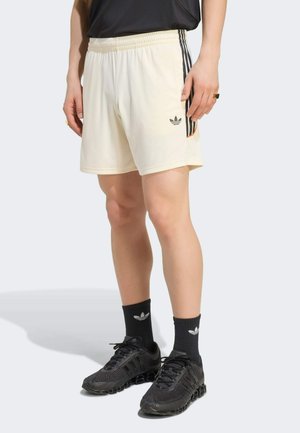 Osoba ubrana w kremowe szorty z czarnymi paskami i logo Adidas, czarne skarpety Adidas oraz czarne sportowe buty stojąca na szarej podłodze.