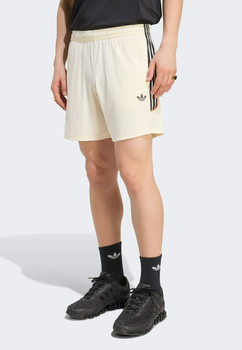 Personne portant un short crème avec des rayures noires et un logo Adidas, des chaussettes Adidas noires et des baskets de sport noires, debout sur un sol gris.