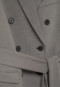 Blazer doppiopetto in tessuto pied de poule bianco e nero, con bottoni neri, una cintura e un tessuto testurizzato.