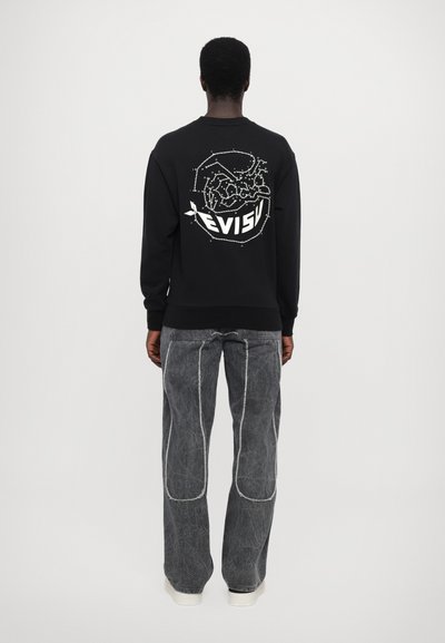 Sweat-shirt noir avec un design graphique blanc et du texte au dos. Jean gris avec des coutures blanches contrastantes. Chaussures simples.