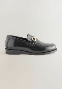 Mocassin en cuir noir à bout rond, avec une boucle décorative en or et des coutures, reposant sur une semelle plate en caoutchouc.