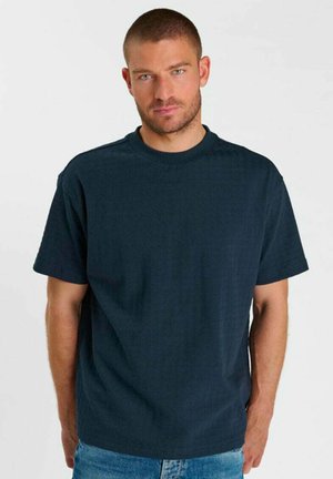 Uomo con capelli corti e barba che indossa una maglietta blu scuro semplice e jeans azzurri, in piedi davanti a uno sfondo bianco.