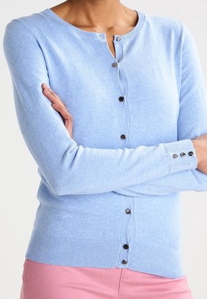 Personne portant un cardigan bleu clair boutonné avec des boutons en métal et un pantalon rose, les bras croisés sur la poitrine.