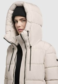 Puffer jacket in light beige, featuring a large quilted hood, zippered front, and black drawstrings. Soft texture with contrasting accents.