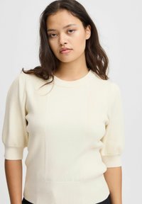 Krēmveida ribota adīta džemperis ar īsām puff piedurknēm, ar vertikālu paneļu izstrādājumu un noapaļotu kakla izgriezumu, izgatavots no mīksta, elastīga materiāla.