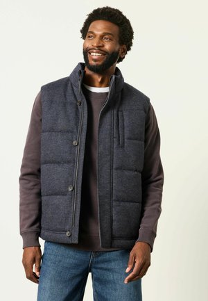 Man smiling and looking right wearing a dark grey quilted vest over a brown sweatshirt and blue jeans against a plain light background.