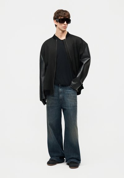 Jeune homme portant une veste bomber noire surdimensionnée aux manches en cuir, des gants noirs, des lunettes de soleil, un jean bleu ample et des chaussures noires, debout.