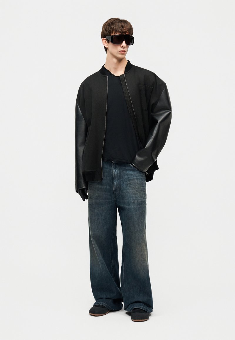 Jonge man die een oversized zwarte bomberjack met leren mouwen draagt, zwarte handschoenen, een zonnebril, losse blauwe jeans en zwarte schoenen, staand.