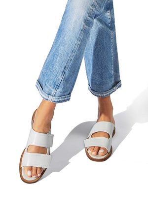 Nærbillede af fødder, der bærer hvide lædersandaler med to stropper, parret med opsmøgede blå jeans på en hvid baggrund.