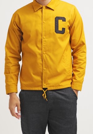 Man draagt een mosterdgele jas met knopen en een zwarte letter "C" op de linkerborst en donkergrijze broek, één hand in de zak.