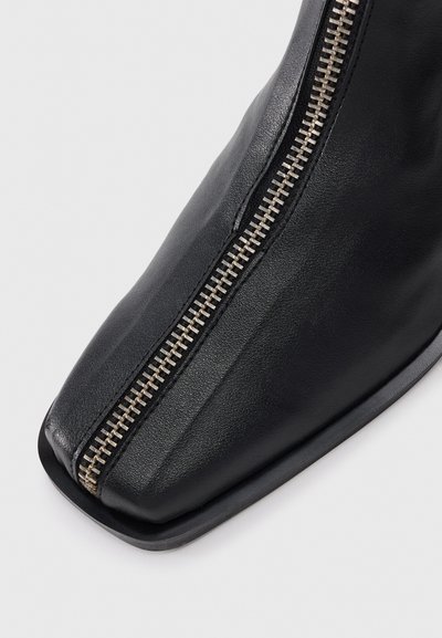 Bota negra de cuero hasta el tobillo con punta cuadrada y detalle de cremallera metálica en el costado, que presenta una textura suave y un diseño minimalista.