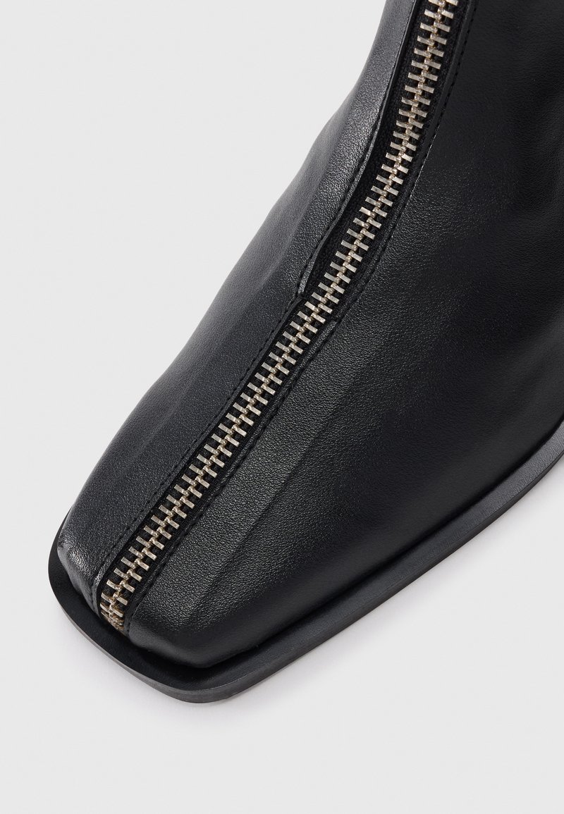 Bottines en cuir noir avec un bout carré et un détail de fermeture éclair métallique sur le côté, affichant une texture lisse et un design minimaliste.
