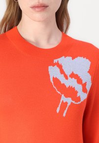 Maglione arancione lavorato a maglia con un design floreale grigio sulla spalla sinistra e un classico colletto rotondo a costine. Tessuto morbido.
