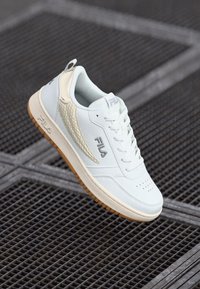 Hvit sneaker med teksturerte detaljer, Fila-logo, vevd design på siden, perforert tå og lys brun yttersåle. Flate snørebånd og polstret krage.