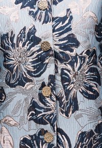 Μπλε ύφασμα με μεγάλο λουλουδάτο μοτίβο σε ναυτικό μπλε και ανοιχτό ροζ, με τέσσερα υφασμένα στρογγυλά χρυσά κουμπιά σε κάθετη σειρά.