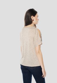 Beige t-shirt met korte mouwen en open schouderontwerp, gecombineerd met donkerblauwe spijkerbroek. Achteraanzicht, minimale details.