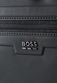 Κοντινό πλάνο μαύρου υφάσματος με υφή, με φερμουάρ και μεταλλικό λογότυπο "BOSS" σε τσάντα ή αξεσουάρ.