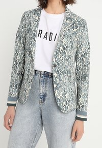 Blazer de estampado de leopardo en azul claro y crema, con un solo botón, solapas de muesca y puños a rayas. Llevado sobre una camiseta gráfica blanca.
