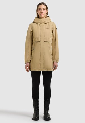 Vrouw draagt een beige parka-jack met capuchon, zwarte legging en zwarte enkellaarsjes, staand tegen een effen grijze achtergrond.
