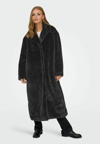 Donna che indossa un lungo cappotto di pelliccia sintetica nera su pantaloni neri e mocassini neri, in piedi davanti a uno sfondo chiaro e semplice.