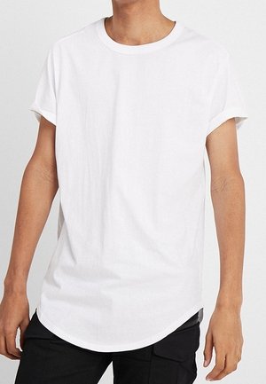 Personne portant un t-shirt blanc uni à manches courtes avec des poignets retroussés et un pantalon noir, se tenant devant un fond uni.