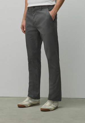 Man wearing dark gray straight-leg pants and white sneakers, standing with one hand in pocket against a plain background.