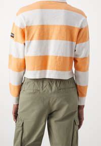 Bauchfreies Langarmoberteil mit orange-weißen Streifen; aus weichem Stoff gefertigt. Kombiniert mit lässigen olivgrünen Cargo-Hosen.