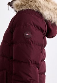 Bordowa kurtka puchowa z futrzanym kapturem, z okrągłą naszywką na lewym ramieniu i pikowaną strukturą.