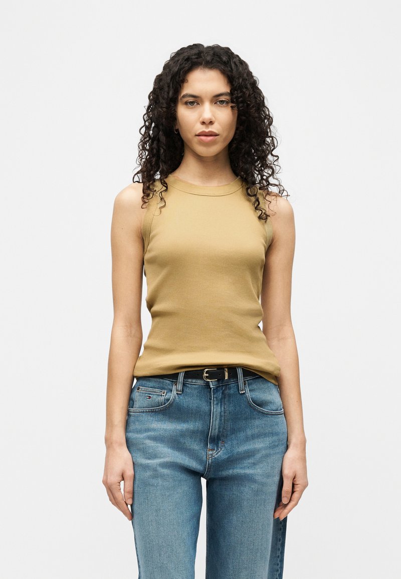 Kobieta o kręconych włosach ubrana w beżową, bezrękawnikową bluzkę i niebieskie jeansy, stojąca na tle jednolitego białego tła.