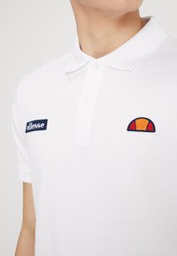 Weißes Poloshirt aus strukturiertem Material. Verfügt über ein dunkelblaues Logo-Patch und ein mehrfarbiges gesticktes Design auf der Brust. Kurze Ärmel.