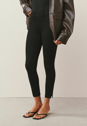 Person wearing black slim-fit ankle pants, black flip-flops, and a dark brown leather jacket with hands in pockets.