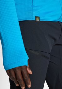 Fazonos kék hosszú ujjú ing textúrált anyagból, hüvelykujjnak kialakított lyukkal, fekete nadrággal párosítva, elegáns cipzáras zsebbel.