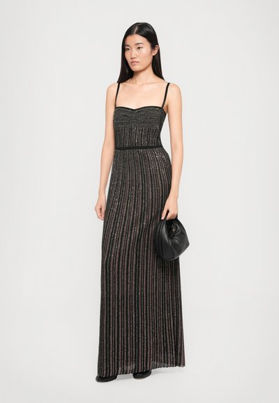 Maxi dress in black with vertical metallic stripes, a sweetheart neckline, thin straps, and a fitted waist. Model holding a black handbag.
