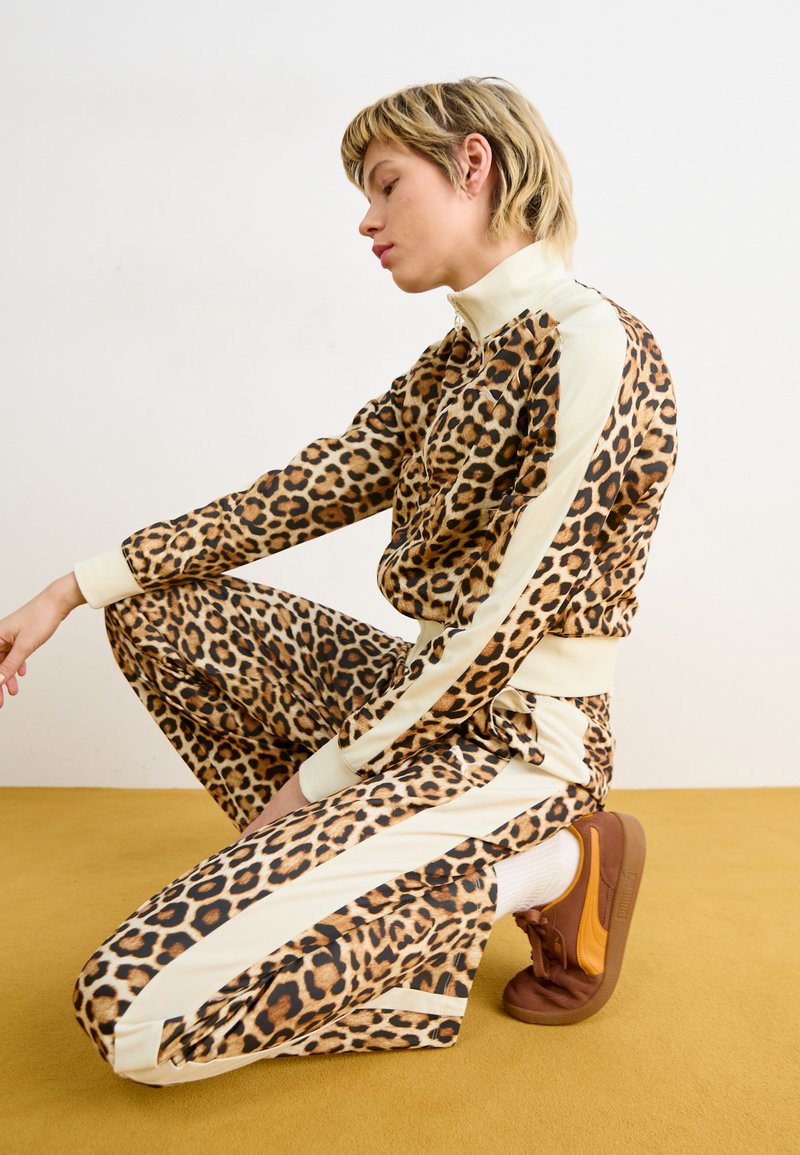 Survêtement en imprimé léopard avec un haut et un pantalon assortis, accents crème et col montant. Des baskets marron complètent la tenue. Fond jaune.