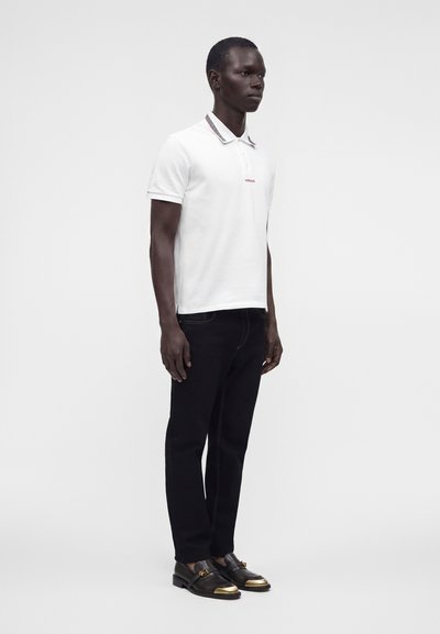 Homme portant un polo blanc avec col à motif, un pantalon noir et des chaussures en cuir noir avec des bouts dorés, se tenant devant un fond uni.