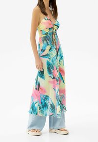 Een vrouw in een mouwloze tropische bloemen-maxidress, gedragen over lichtblauwe wijde jeans en witte platform sandalen.